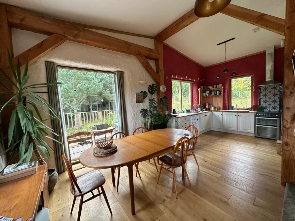 A spacious kitchen and dining area with wooden beams, a wooden table and chairs, white cabinets, large windows, and houseplants—an inviting space showcasing eco-building principles and overlooking a deck and trees outside.